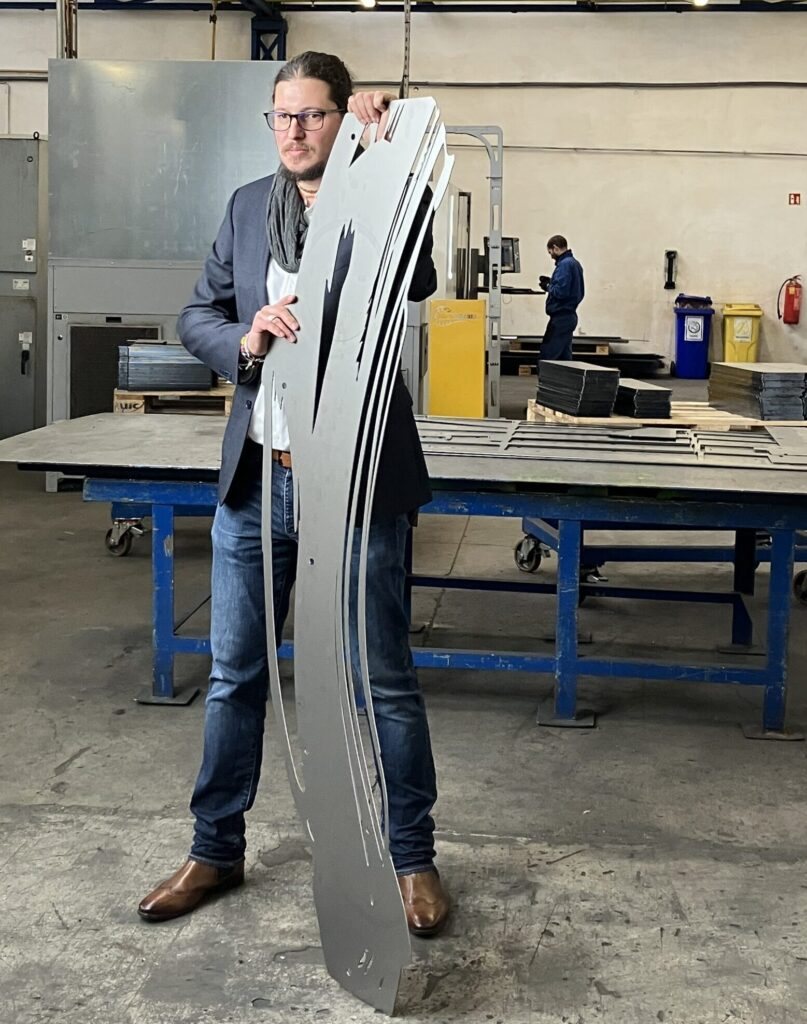 Péter Alasztics holding a tall laser-cut steel artwork in the Intertechnika workshop