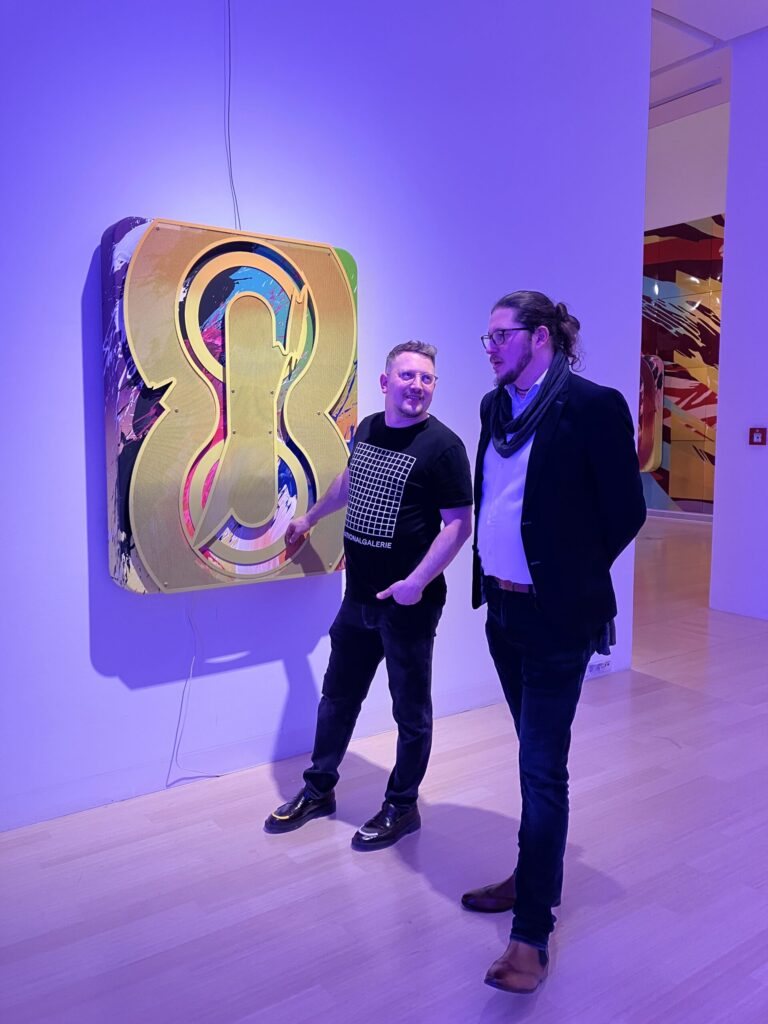 Márton Nemes and Péter Alasztics in front of a colourful metal artwork under purple light