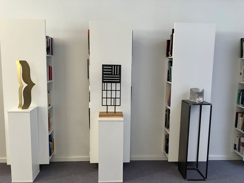 Three metal sculptures on white and black plinths between bookshelves at the Liszt Institute