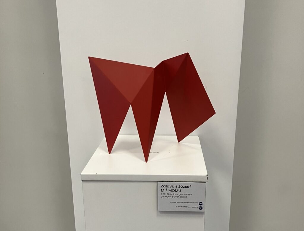 Red folded steel sculpture shaped like the letter M on a white plinth