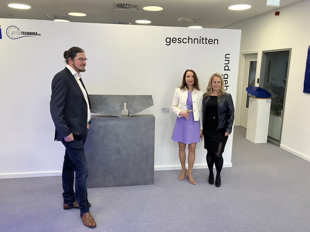Three people standing at the opening of the “Cut and Bent” exhibition next to a steel sculpture