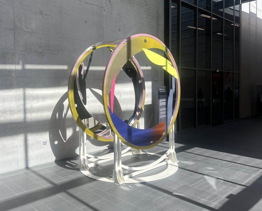 Circular sculpture “Superposed” with coloured metal rings in a sunlit concrete hall